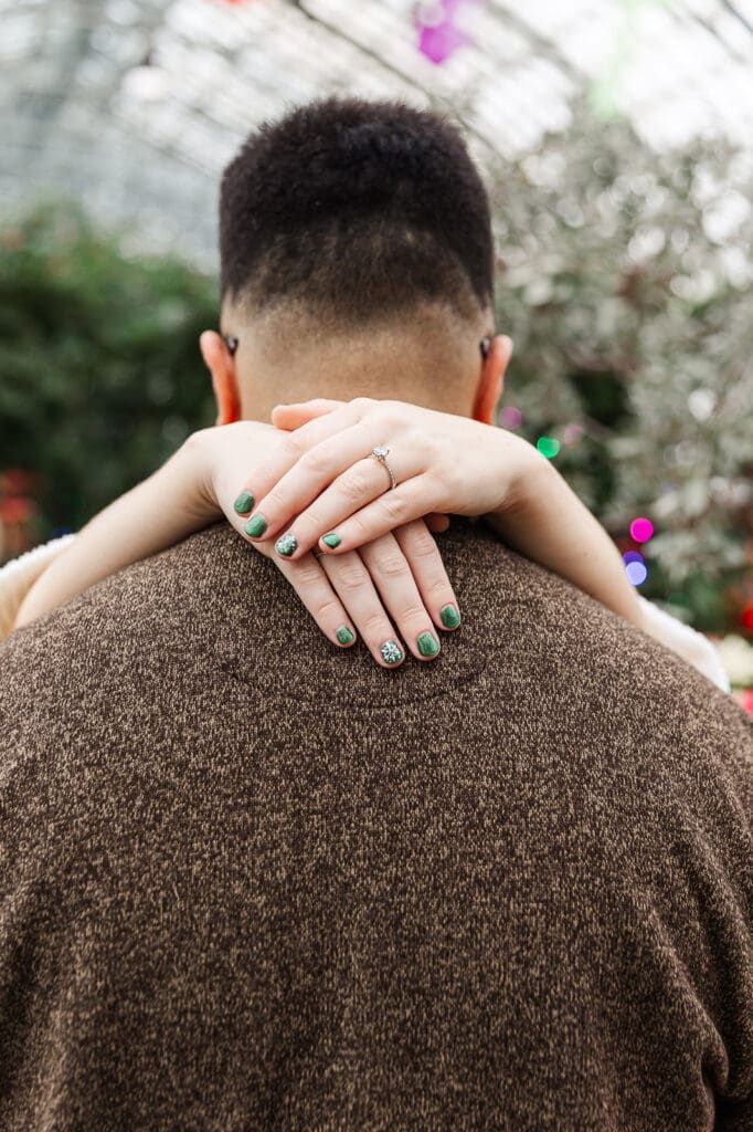 The brides' hands wrapped around the groom's neck showcasing her green nails themed for their Christmas engagement session that focuses on her engagement ring