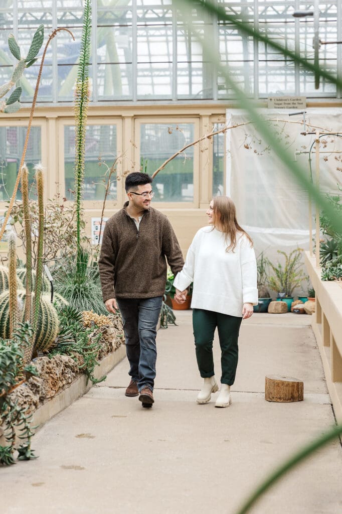 Couple laughing and walking hand-in-hand through a bright dessert-themed room during their Garfield Park Conservatory engagement session.